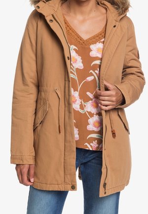 Bruine hoodie parka met nepbont, voorzien van zijzakken en een trekkoord in de taille, gelaagd over een bruin top met bloemmotief.