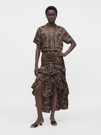 Γυναίκα που στέκεται φορώντας καφέ crop top με paisley μοτίβο και κοντά μανίκια και ασορτί ασύμμετρη φούστα παρεό, φορώντας μαύρα πέδιλα με τακούνι και ανοιχτά δάχτυλα.