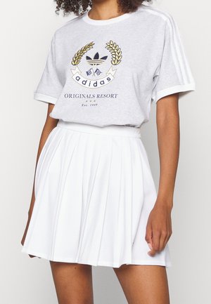 Kobieta ubrana w szarą koszulkę Adidas Originals Resort z złoto-granatowym logo oraz białą plisowaną spódnicę.