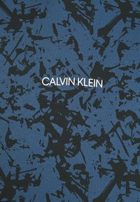 Tissu bleu avec motif éclaboussé abstrait noir et texte blanc « Calvin Klein » centré.