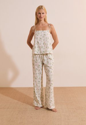 Femme debout pieds nus sur un sol beige, portant un ensemble pyjama en soie blanche avec un petit motif floral vert et rose, les mains derrière le dos.