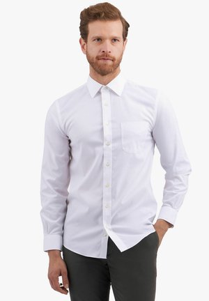 Man draagt een wit overhemd met lange mouwen en een borstzak, staat met één hand in zijn zak tegen een effen achtergrond.