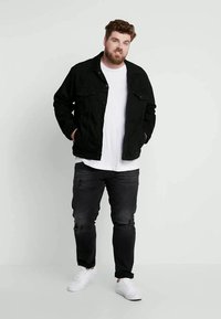 Homem com barba em pé, vestindo uma jaqueta preta, t-shirt branca, jeans pretos e ténis brancos contra um fundo claro e simples.