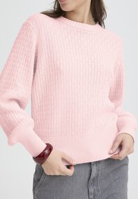 Pull en maille côtelée rose avec manches longues et détails texturés, doté d'une ceinture large et porté par un mannequin portant un bracelet rouge.