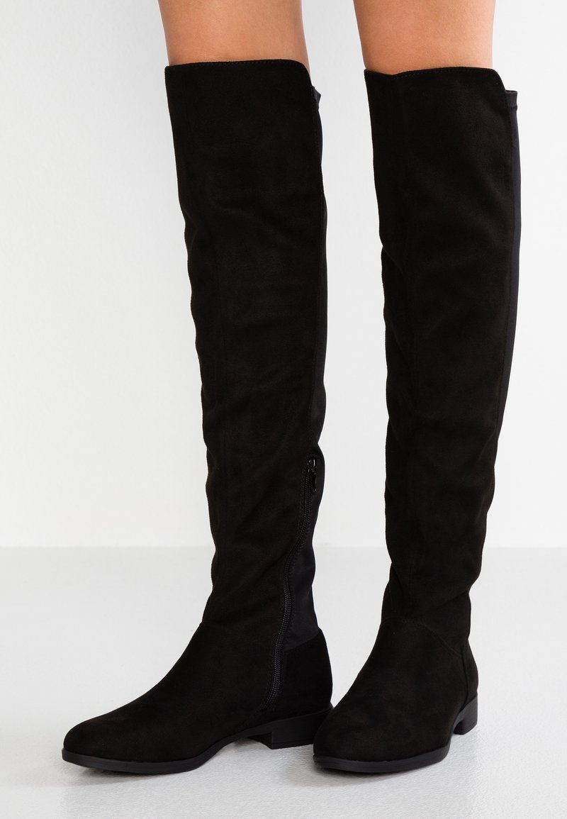 Bottes plates noires jusqu'aux genoux avec fermeture éclair latérale portées sur des jambes nues, présentées sur un fond clair uni.
