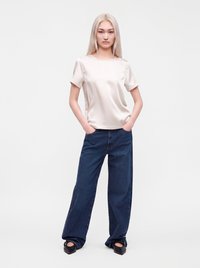 Jeune femme aux longs cheveux blonds, debout, les mains dans les poches, portant un top crème en satin à manches courtes, un jean bleu à jambes larges et des chaussures noires.