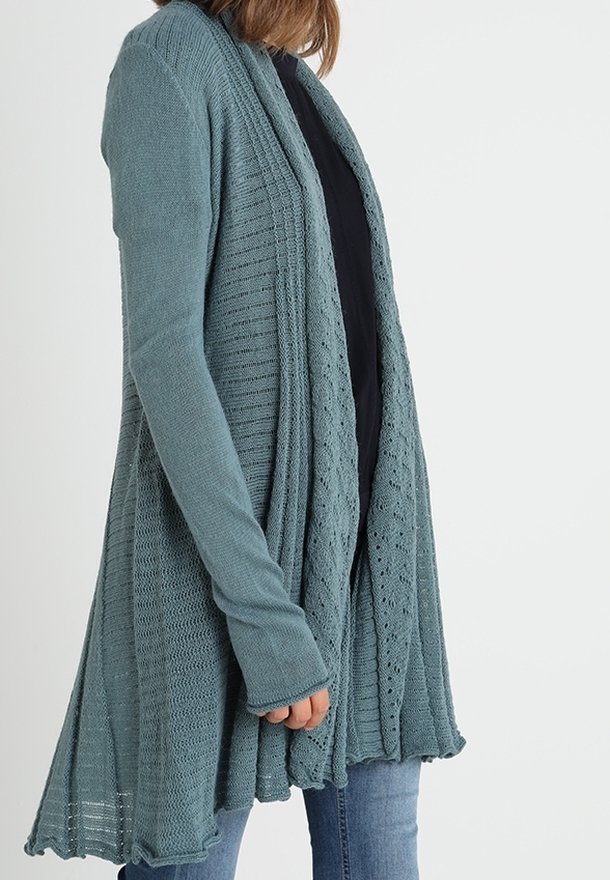 Kvinna iförd en tealfärgad stickad cardigan med öppen front och texturerade mönster över en svart topp och blå jeans, ståendes mot en enfärgad vit bakgrund.