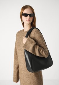 Black leather shoulder bag with a curved shape and smooth texture, paired with a brown knitted sweater featuring a wide, ribbed cuff.