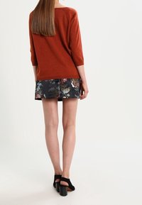 Femme portant un pull couleur rouille, une mini-jupe à motifs floraux et des sandales à talons carrés noires, debout de dos sur fond blanc.