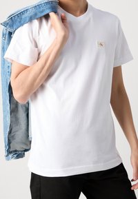 Persona che indossa una t-shirt bianca Calvin Klein e pantaloni neri, tenendo una giacca di jeans blu sulla spalla, su uno sfondo semplice.