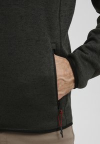 Veste en polaire zippée gris foncé avec une finition texturée, mettant en avant une main dans une poche latérale et un détail à cordon au niveau de l'ourlet.