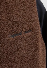 Nahaufnahme eines braunen, strukturierten Fleece-Stoffs mit dunkel gesticktem, kursivem Text auf schwarzer Kleidung darunter.