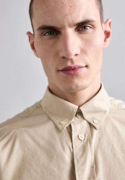 Beige bomulds skjorte med button-down krave, der har en glat tekstur og to knapper foran til lukning. Nærbillede.