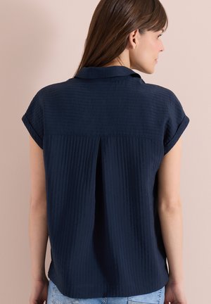 Femme de profil portant un chemisier texturé bleu marine à manches courtes avec un pli au dos et un jean bleu clair.