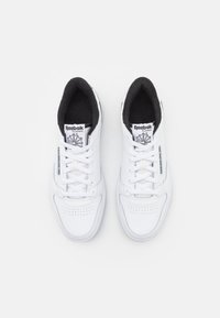 Bílé syntetické tenisky s perforovanou špičkou, plochými tkaničkami a černým vnitřním podšívkou. Obsahují branding Reebok na jazyku a na bocích.