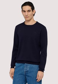 Marineblauer Pullover mit Rundhalsausschnitt aus glattem Stoff mit gerippten Bündchen und Saum, getragen über hellblauen Jeans. Dezentes Logodetail auf der Brust.