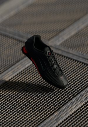 Scarpa sportiva nera con dettagli rossi e logo Nike, appoggiata su una superficie di griglia metallica strutturata con ombre diagonali.