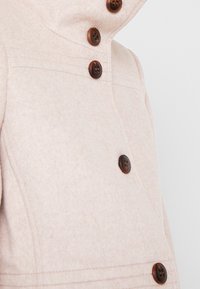 Manteau en laine rose clair avec un col montant, comprenant quatre boutons marron foncé et des coutures discrètes. Texture lisse et coupe ajustée.