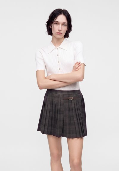 Jeune femme aux cheveux courts et foncés, portant une chemise blanche à manches courtes boutonnée et une jupe plissée grise avec un petit détail ceinture.