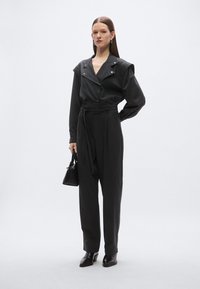 Črni kombinezon z širokim ovratnikom, gumbim spredaj in pasom okrog pasu. Stiliziran z majhno črno torbico in elegantnimi črnimi škornji.