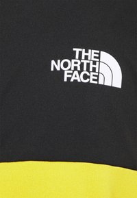 Czarny sweter z pionowym żółtym paskiem na dolnym końcu. Posiada białe logo "The North Face" na klatce piersiowej. Gładka faktura materiału.