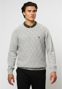 Šviesiai pilkas megztinis su deimanto raštu, apvaliu kaklu ir raukšlėmis ant rankovių. Priekyje yra maža tamsi logotipas. Sujungtas su juodomis kelnėmis.