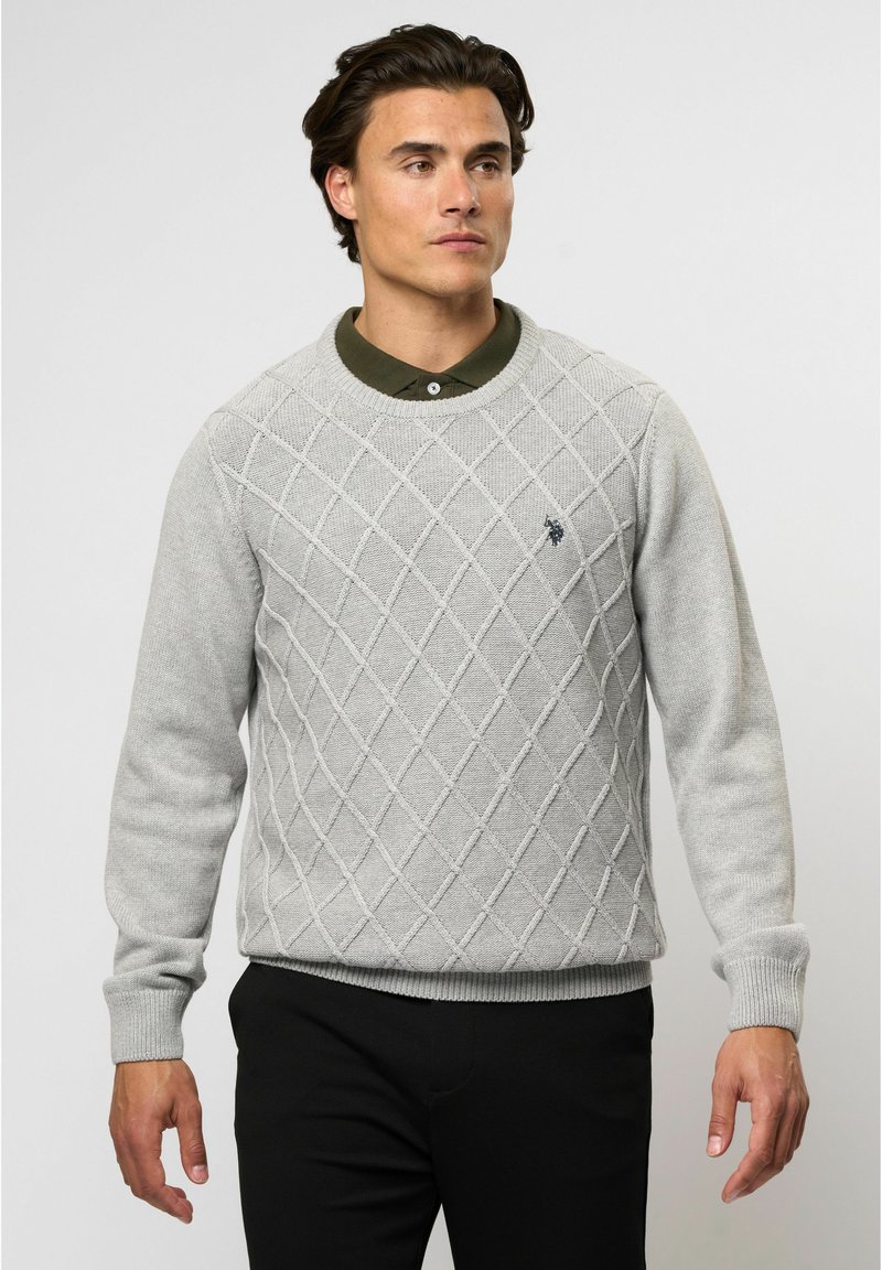 Šviesiai pilkas megztinis su deimanto raštu, apvaliu kaklu ir raukšlėmis ant rankovių. Priekyje yra maža tamsi logotipas. Sujungtas su juodomis kelnėmis.