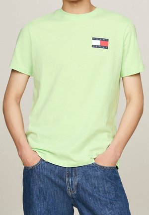 Personne portant un T-shirt vert clair à manches courtes avec le logo Tommy Jeans sur la poitrine et un jean bleu en denim, les mains dans les poches.