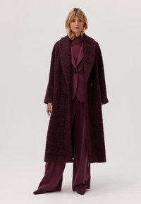 Bordowy długi płaszcz o teksturowanej, puszystej powierzchni, z szerokimi klapami i zapięciem na jeden guzik, noszony na zestawie pasującym do garnituru.
