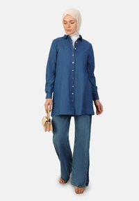 Camisa tunic de mezclilla en azul oscuro con un cierre de botones al frente, mangas largas y un corte relajado, combinada con jeans de pierna ancha y un bolso beige claro.
