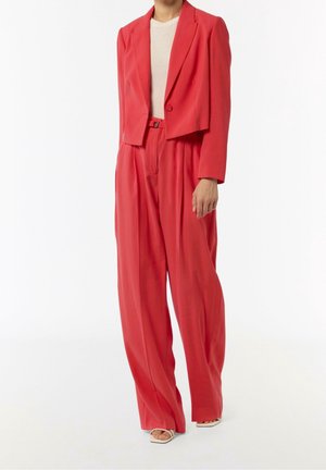 Frau trägt locker sitzendes, rotes kurzes Blazer, passende hoch taillierte, weit geschnittene Hosen, weißes Oberteil und weiße offene Sandaletten auf weißem Hintergrund.