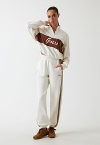 Crème zip-up sweatshirt met een bruine panel en het 'Guess' logo, gecombineerd met crème sweatpants met bruine zijstrepen en logo. Bruine schoenen.