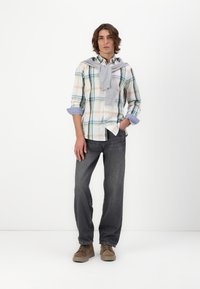 Chemise à manches longues à carreaux blancs, verts et oranges avec des poignets bleus retroussés, associée à un jean gris et des chaussures beiges, et un pull gris drapé sur les épaules.