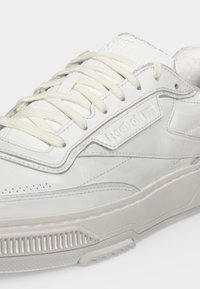 Λευκό δερμάτινο αθλητικό παπούτσι Reebok με κορδόνια, ανάγλυφο λογότυπο στο πλάι και υφασμένο ελαστικό πέλμα ορατό σε κοντινή λήψη.