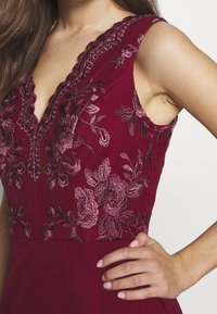 Burgunderfärgad klänning med djup V-ringning, bluma brodyr i rosa nyanser och en åtsittande överdel med en slät yta.