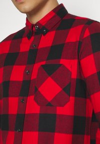 Camisa de cuadros en rojo y negro con cuello abotonado, que presenta botones negros y un bolsillo en ángulo en el lado izquierdo del pecho. Textura de tela suave.