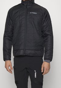Czarna pikowana kurtka z wysokim kołnierzem, zamkiem błyskawicznym i logiem Terrex. Charakteryzuje się gładką fakturą i strukturalnym wzorem. Nosi się ją na czarnych spodniach.