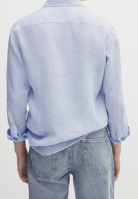 Lichtblauw, lang-geleed, linnen shirt met opgerolde mouwen, gedragen over medium-wash denim jeans. De achterkant toont een eenvoudig ontwerp en textuur van de stof.