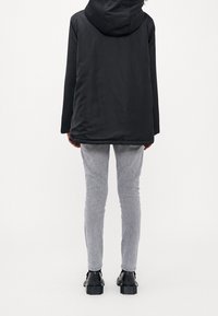 Osoba stojící zády, oblečená v černé kapucňové bundě, světlých šedých úzkých kalhotách a černých botách s hrubou podrážkou, na bílém pozadí.