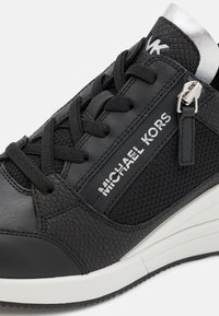 Sort sneaker med en tekstureret læderfinish, mesh-paneer, tykke snørebånd og en sølv akcent ved anklen. Logo vist på siden.