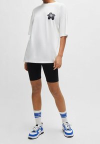 Biała bawełniana koszulka z krótkimi rękawami i czarnym kwiatowym motywem na piersi, zestawiona z czarnymi krótkimi spodenkami rowerowymi i niebiesko zaakcentowanymi sneakersami.