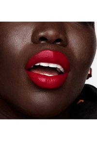 Couleur à lèvres rouge vif sur des lèvres pleines, mettant en valeur une finition brillante. Le teint foncé contraste avec cette teinte vibrante, soulignant la texture et la forme.