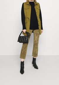Femme portant une veste sans manches vert olive, un haut noir à manches longues, un pantalon court en daim vert olive, des bottines noires, tenant un sac à main noir matelassé.