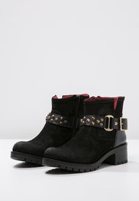 Black suede ankle boots with a side zipper, gold studs on a black belt, and a red interior lining. Chunky heel and textured sole.