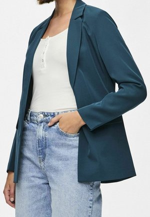 Vrouw draagt een teal blazer over een wit geribd henley-topje met blauwe high-waisted jeans, hand in zak, tegen een effen achtergrond.