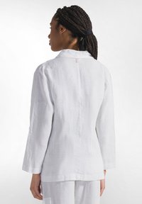 Biała lniana bluzka z długim rękawem i kołnierzykiem, o prostym kroju i lekko luźnym fasonie. Widoczny szew z tyłu.