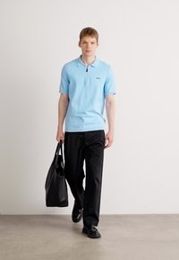 Jovem a caminhar, vestindo uma camisola polo azul clara e calças pretas, a segurar uma grande mala de mão preta na mão direita, fundo branco liso.