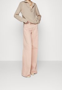 Persoon in een beige lange mouwen top en lichte roze wijde broek, staand met handen in de zakken en tatoeage zichtbaar op de pols.