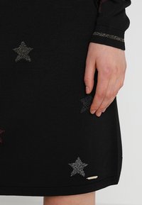 Abito nero a maglia con tessuto texturizzato, caratterizzato da abbellimenti colorati a forma di stella in metallo. Mano parzialmente visibile vicino all'orlo. Dettaglio in hardware argentato.