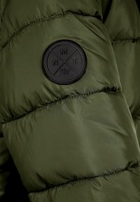 Section de doudoune vert olive avec texture matelassée et un patch en caoutchouc noir circulaire présentant un emblème avec les lettres NMT et le numéro 97.
